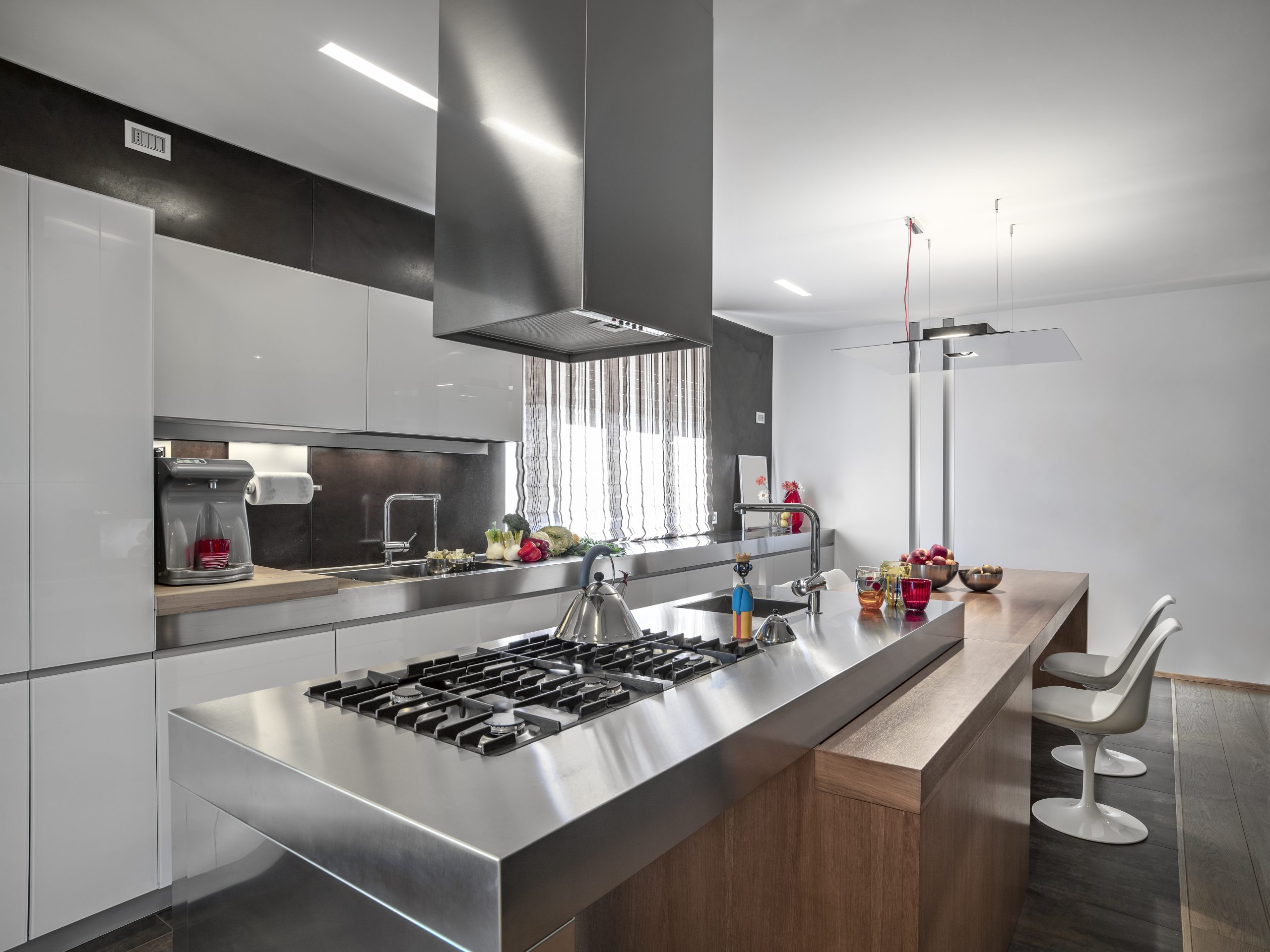 A modern kitchen with a lot of whites and stainless steel. At the forefront is a stainless steel stove with a water pot on it.