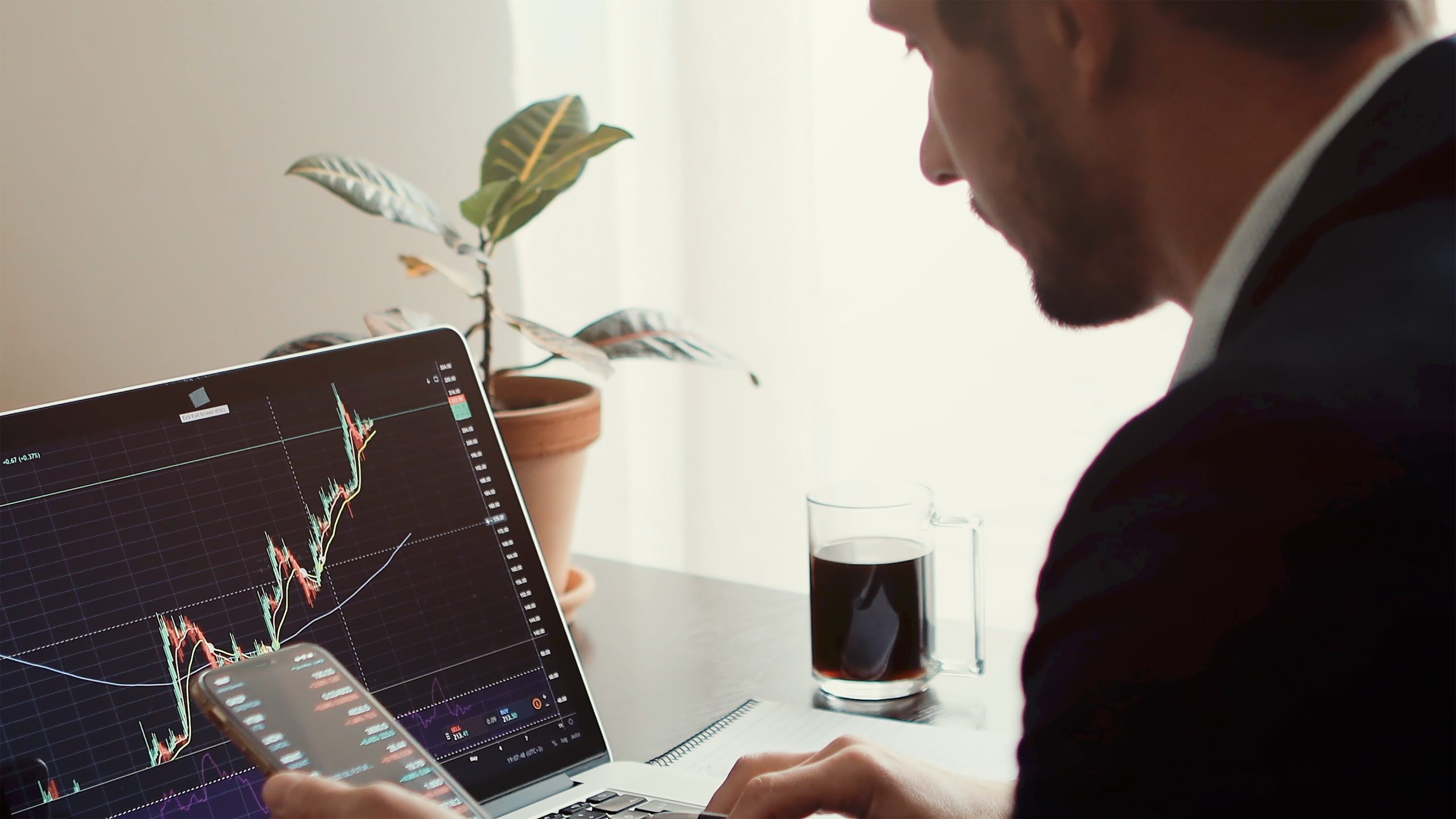 A man in a suit sitting in front of what appears to be a stock chart that shows stocks going up.